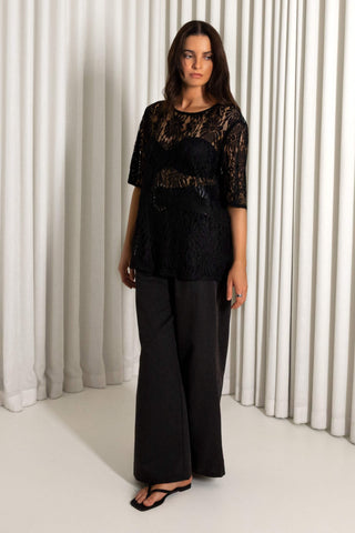 A woman modeling a black lace short-sleeve top with a sheer mesh overlay and black wide-leg pants, standing in front of white pleated curtains.