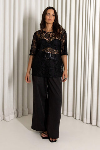 A woman modeling a black lace top with sheer sleeves and wide-leg black pants, standing in front of a white pleated curtain backdrop.
